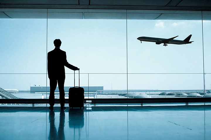 Επιβάτης με βαλίτσ στέκεται όρθιος σε αίθουσα αναμονής στο αεροδρόμιο βλέποντας ένα αεροπλάνο να απογειώνεται.