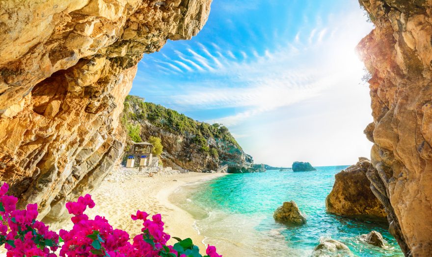 Παραλία με κρυστάλλινα πράσινα νερά ανάμεσα σε βράχους και ροζ λουλούδια.